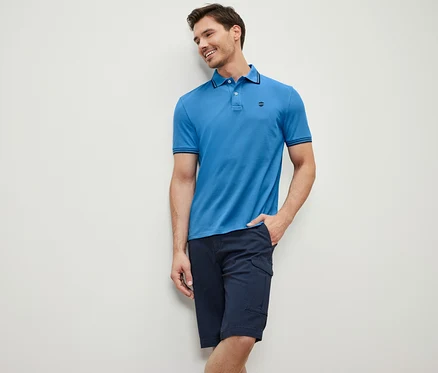 Mann an eine Wand gelehnt trägt ein blaues Piqué-Poloshirt und eine dunkelblaue Shorts.