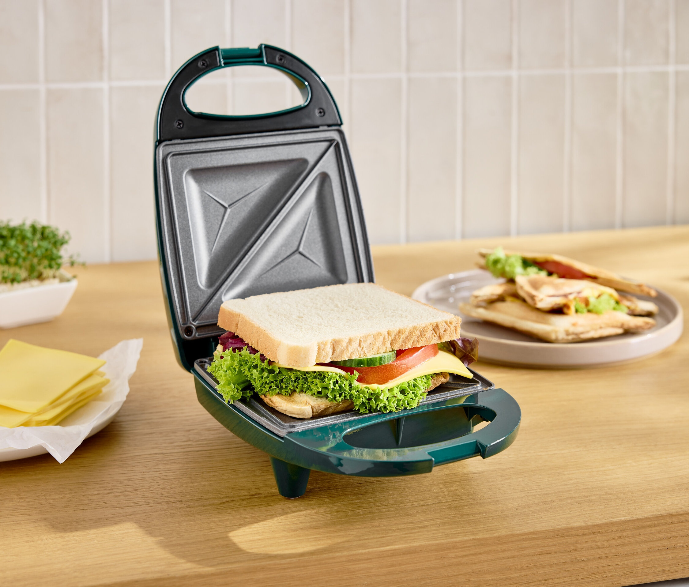 Grüner Sandwichmaker offen mit Toast darin, daneben Teller mit fertigen Toasts und Käse.