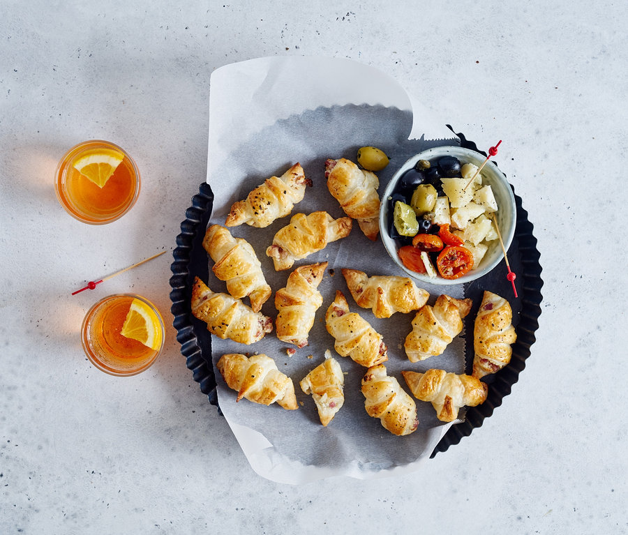 Croissants und eine Schüssel mit Oliven und Tomaten auf einem Backblech. Zwei Getränke mit Orangenscheiben im Hintergrund.