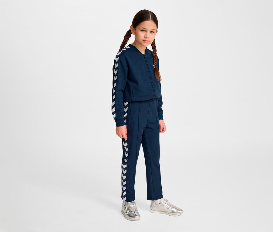 Mädchen mit Zöpfen in marineblauem Hummel HMLJR Archive Regular Poly Pants Anzug und silbernen Schuhen steht vor hellem Hintergrund.