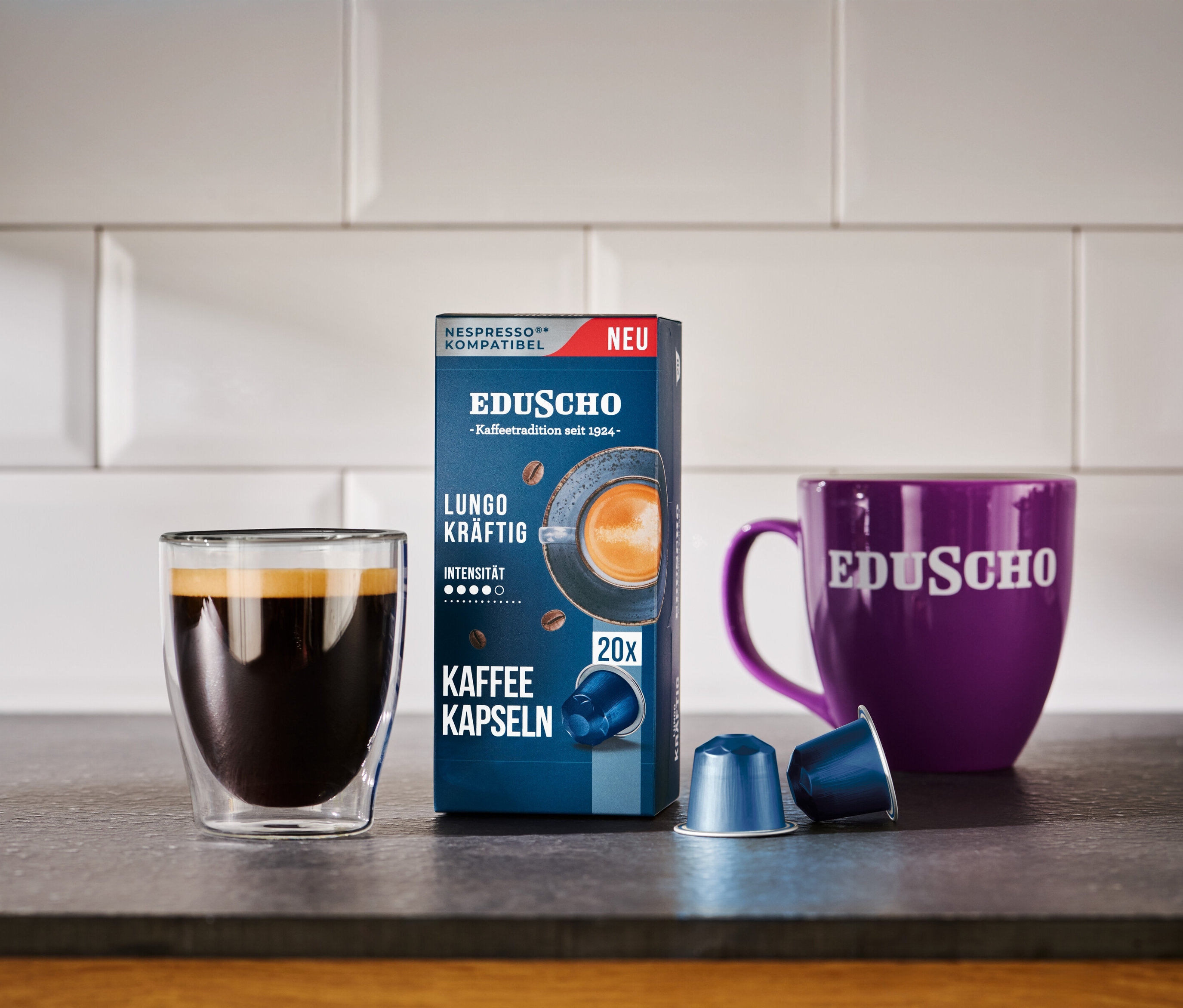 Ein Glas Kaffee neben einer Packung Eduscho Lungo Kräftig Kapseln und einer lila Eduscho Tasse.