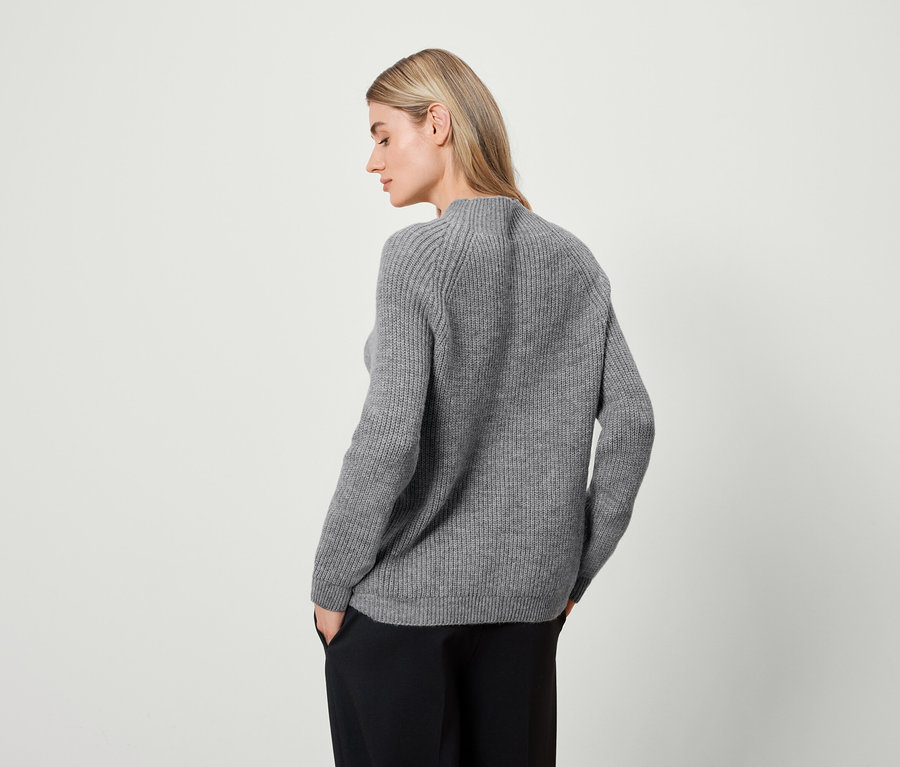 Blonde Frau in grauem Pullover und schwarzer Hose steht mit dem Rücken.