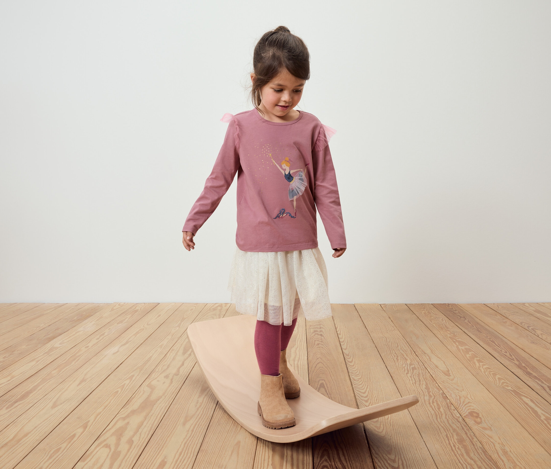 Ein kleines Mädchen steht auf einem Balance Board. Sie trägt ein rosa Langarmshirt mit einem Ballett-Aufdruck, einen weißen Rock und rosa Leggings.