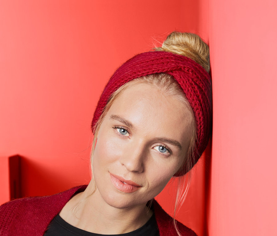 Blonde Frau lehnt an korallenroter Wand und trägt ein Strickstirnband.