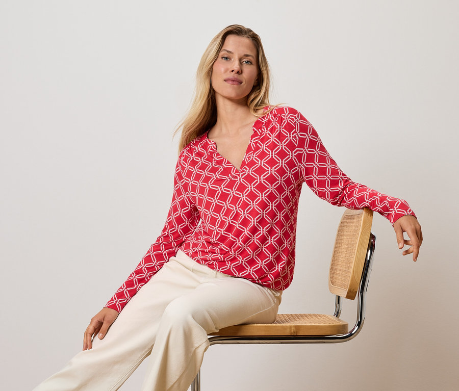 Blonde Frau sitzt auf einem Stuhl, trägt ein rot-weißes Oberteil und cremefarbene Hosen.