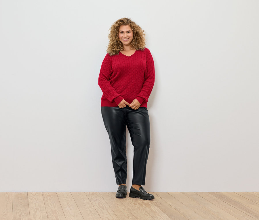 Eine Frau mit lockigem Haar steht vor einer weißen Wand und trägt einen roten Zopfmusterpullover, schwarze Lederhose und Loafer.