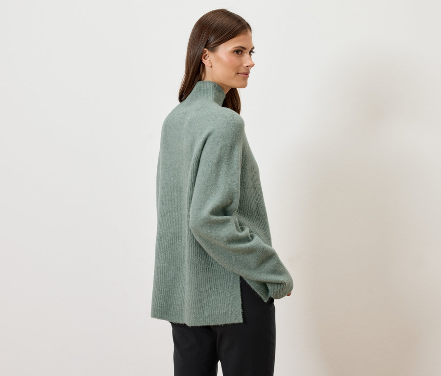 Frau blickt über ihre Schulter und trägt einen grünen Rollkragenpullover und eine schwarze Hose.