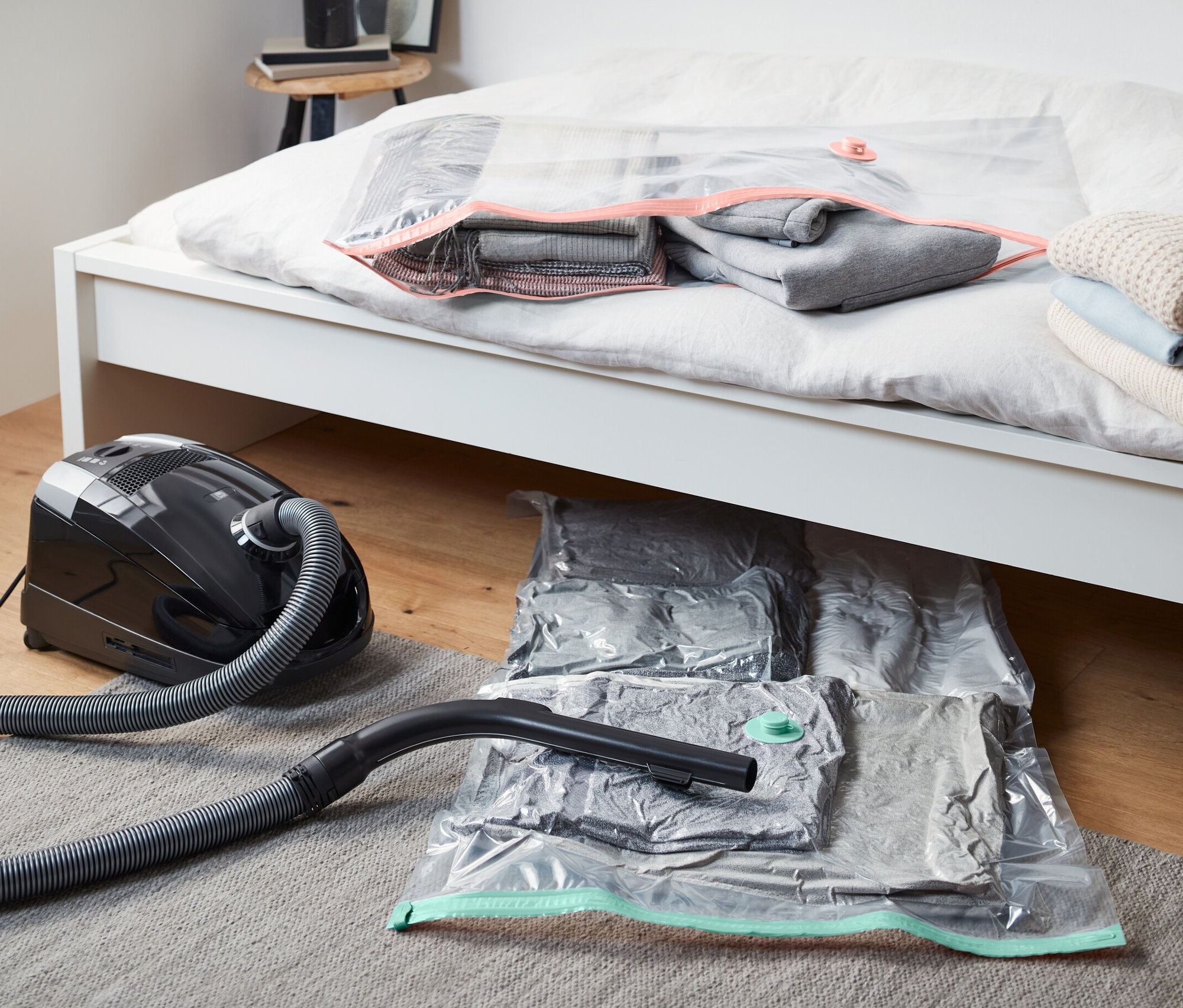 Ein Staubsauger liegt neben zwei Vakuum-Aufbewahrungsbeuteln unter dem Bett. Ein Vakuum-Aufbewahrungsbeutel liegt auf dem Bett.