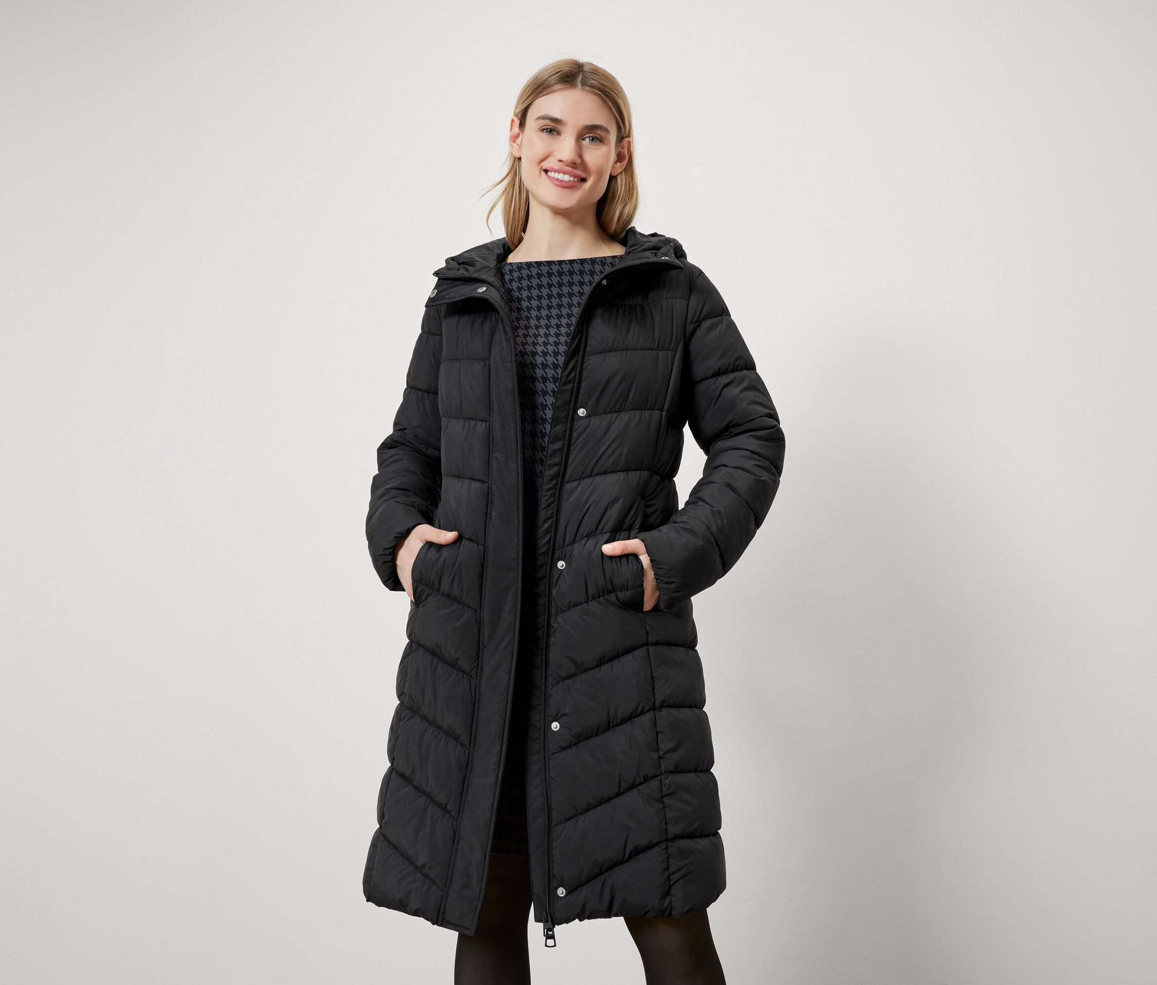 Eine Frau mit blonden Haaren steht und trägt einen langen, schwarzen Steppmantel mit Kapuze.