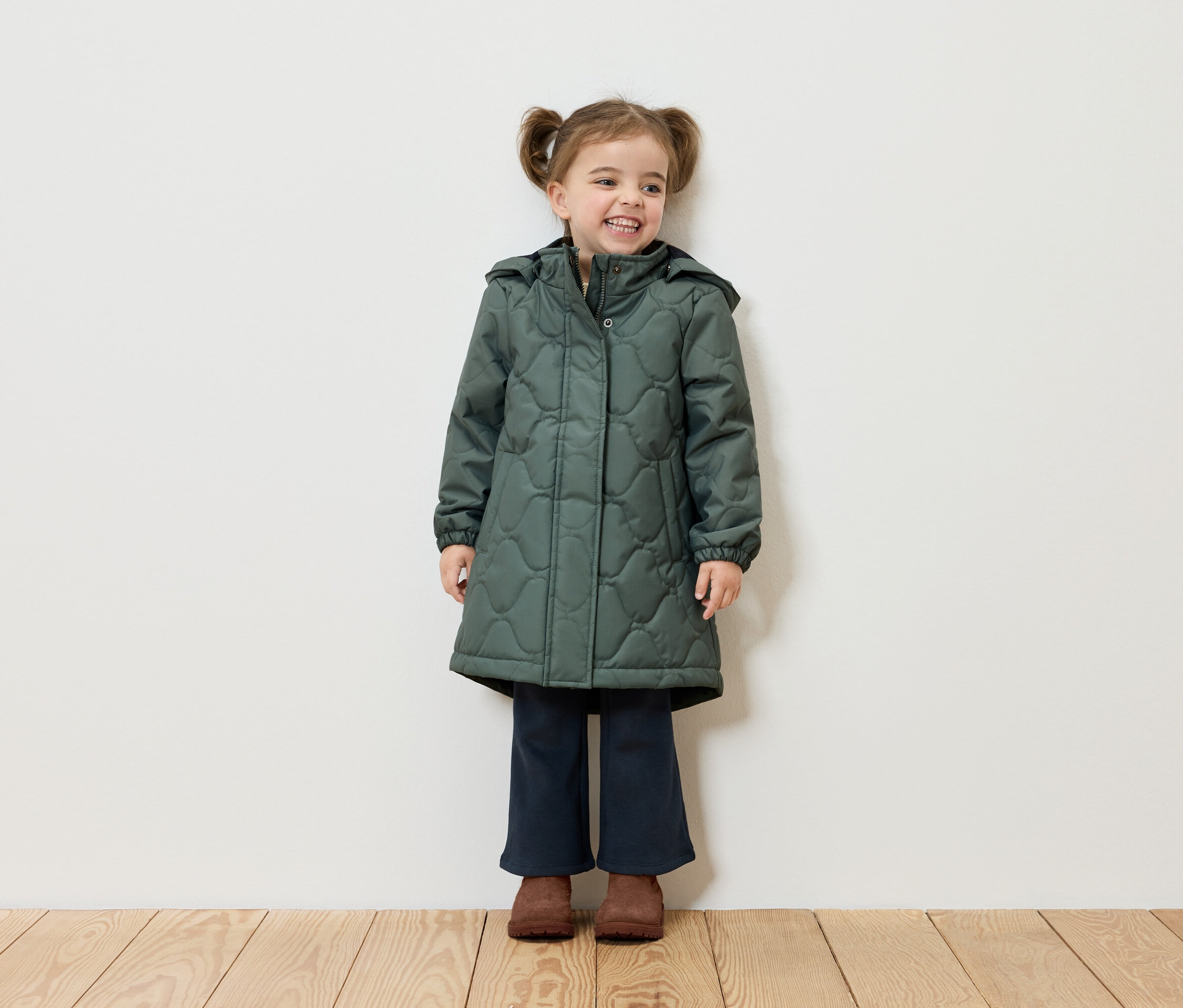 Ein lächelndes Mädchen mit Zöpfen steht vor einer weißen Wand und trägt einen grünen Mantel, dunkelblaue Hosen und braune Schuhe.