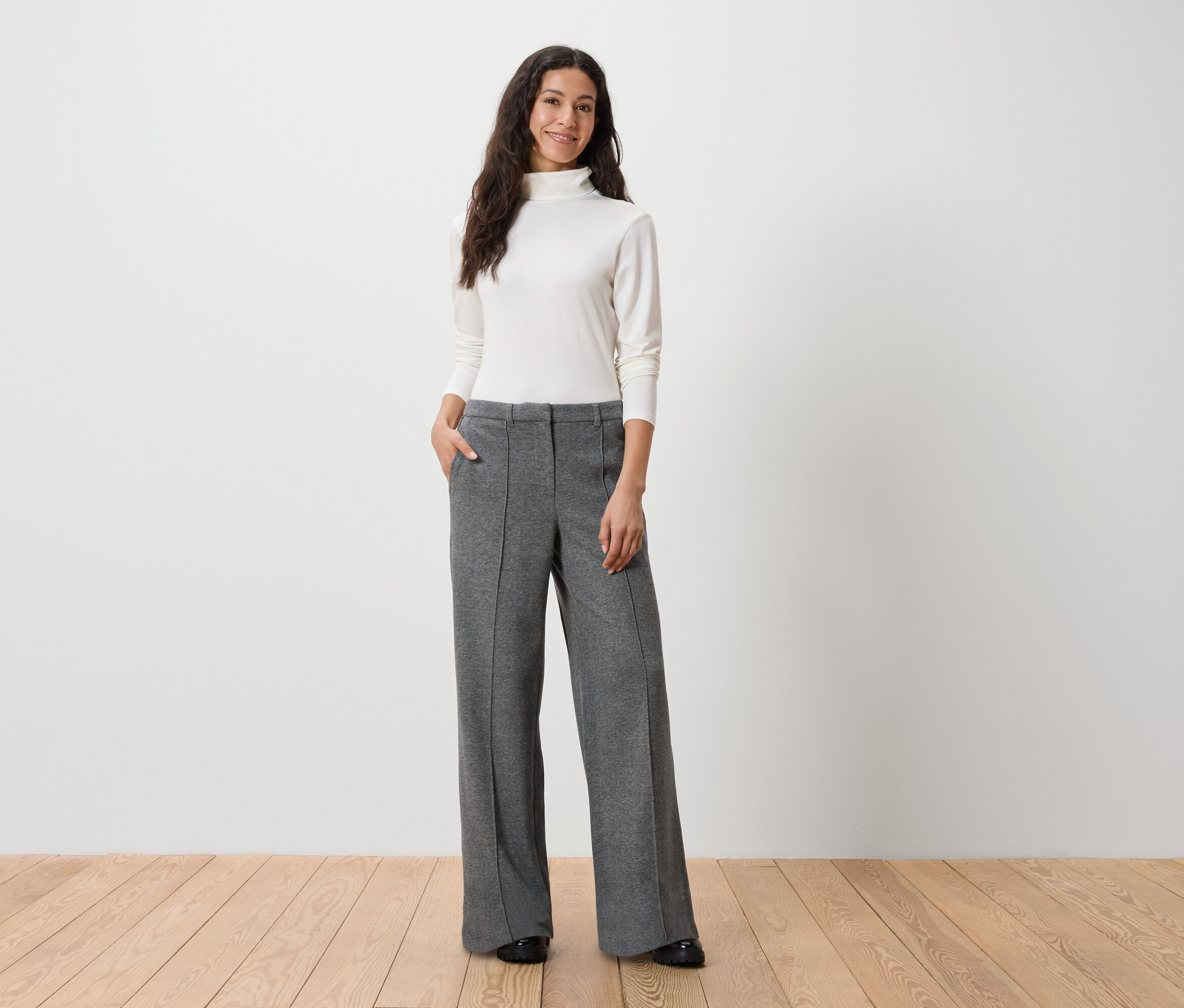 Frau steht mit Händen in den Taschen grauer Hosen und trägt einen weißen Rollkragenpullover.