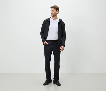 Ein Mann steht und schaut zur Seite, er trägt eine schwarze Hose, ein weißes T-Shirt und eine schwarze Kapuzenjacke.