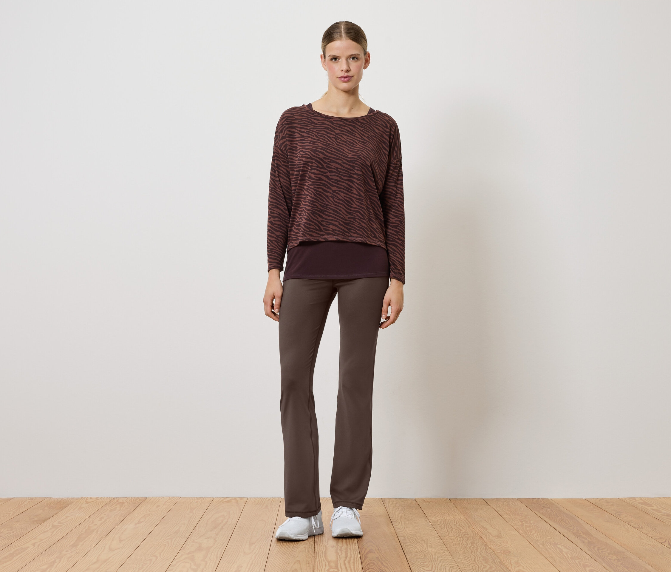 Eine Frau in einem braunen Pullover und braunen Hosen steht auf einem Holzboden.