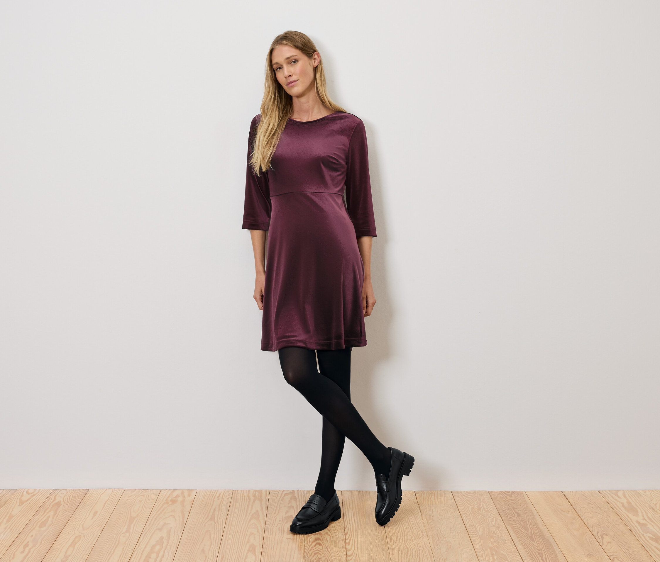 Frau posiert in einem burgunderroten Samtkleid mit Dreiviertelärmeln, schwarzen Strumpfhosen und schwarzen Schuhen.