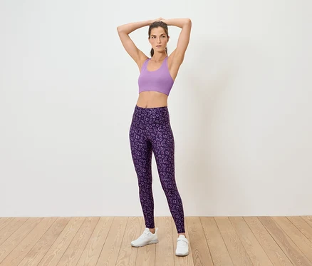 Frau posiert in lila Sport-BH und lila Leggings auf dem Boden.