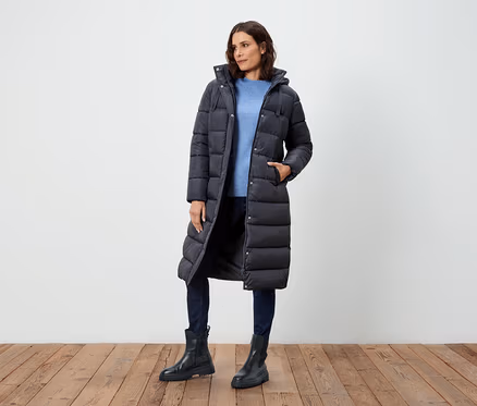 Frau posiert in einem langen, schwarzen Daunenmantel, blauem Pullover, Jeans und schwarzen Stiefeln.