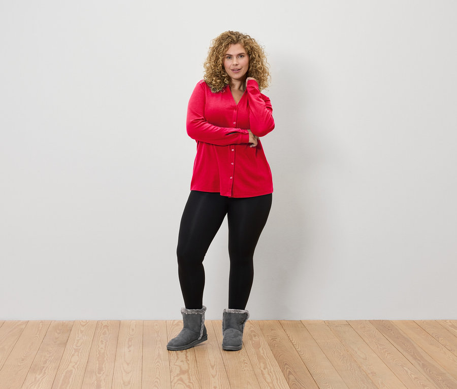 Eine Frau mit lockigem Haar posiert in einer roten Bluse, schwarzen Leggings und grauen Stiefeln vor einer weißen Wand.