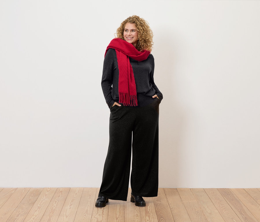 Frau mit lockigem Haar steht mit den Händen in den Taschen. Sie trägt einen roten Schal, ein graues Oberteil und eine schwarze Hose.