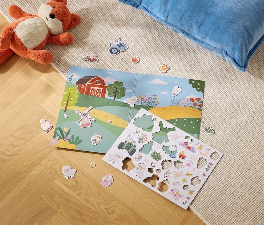 Ein Stofftierfuchs liegt neben einem geöffneten Buch mit einer Bauernhofillustration und einem Bogen mit Bauernhofaufklebern auf einem Teppichboden.