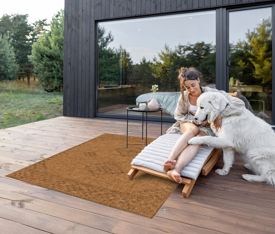 Eine Frau sitzt auf einer Liege auf einer Terrasse und schmust mit einem großen weißen Hund. Auf der Terrasse liegt ein brauner Teppich »Roy«, Antik, ca. 120 x 170 cm.