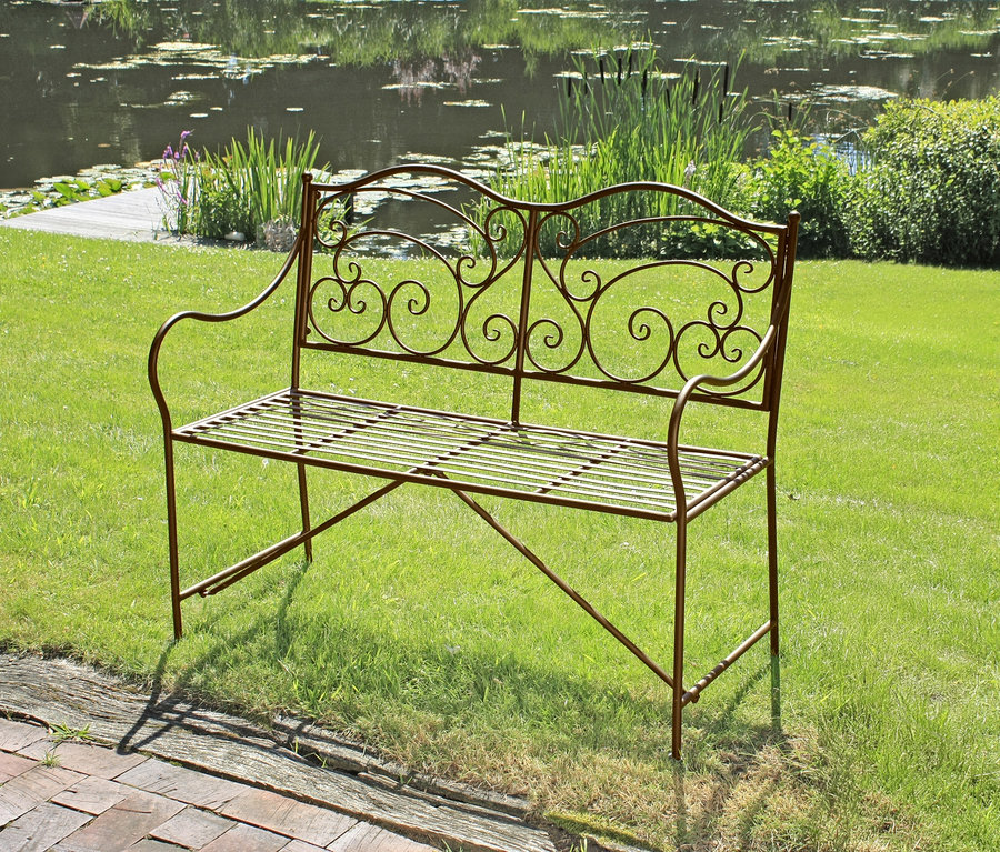 Eine dekorative, bronzefarbene Metallbank mit geschwungenen Verzierungen steht auf einer grünen Wiese vor einem Teich.