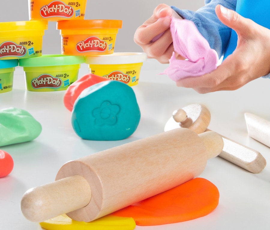 Hände formen rosa Play-Doh. Verschiedenfarbige Play-Doh-Dosen und -Formen sowie ein Nudelholz liegen auf einem Tisch.