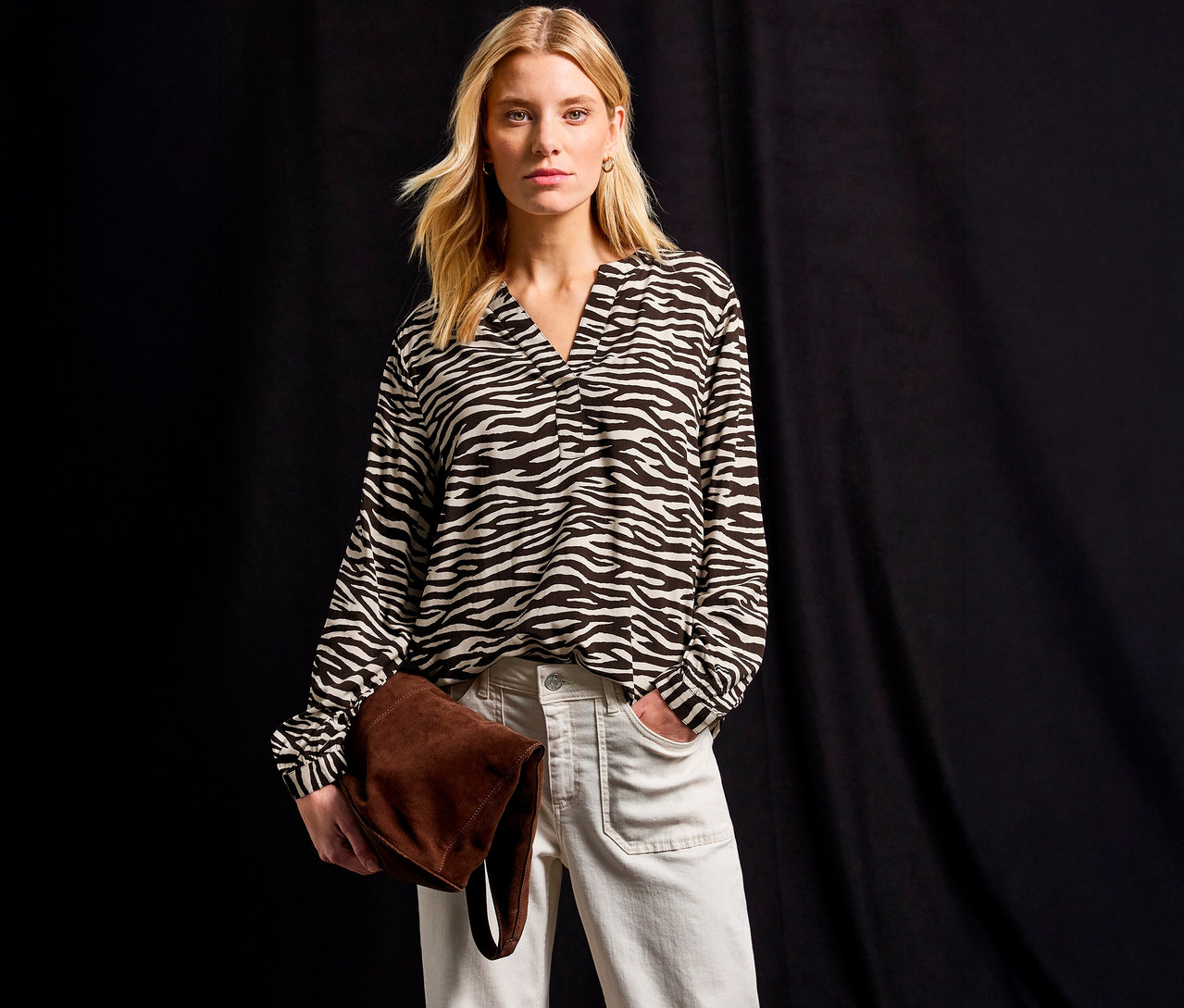 Frau trägt eine Street One Bluse mit Zebramuster, helle Jeans und hält eine braune Tasche vor einem schwarzen Hintergrund.