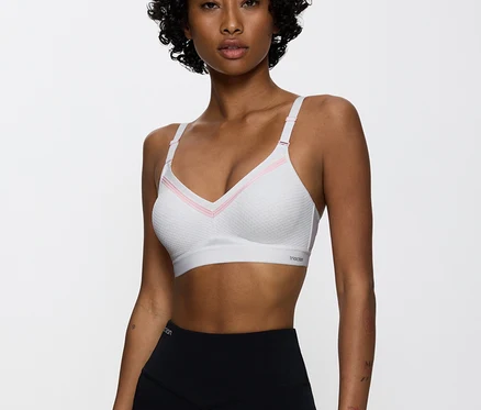 Frau mit lockigem Haar trägt einen weißen TRIUMPH Sport-BH Triaction Free Motion N EX mit rosa Akzenten und schwarze Leggings, blickt nach vorne.