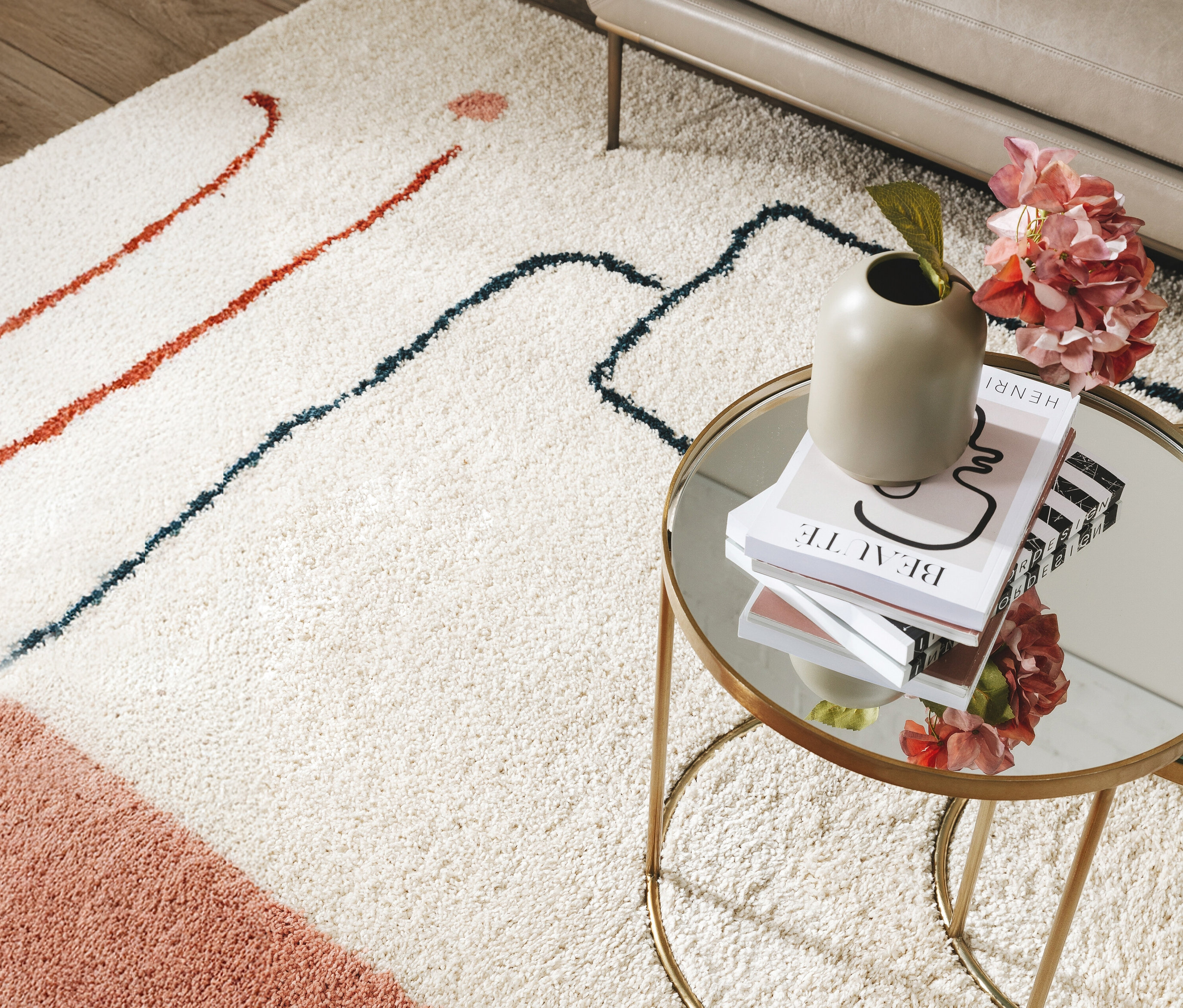 Ein cremefarbener Teppich mit roten und blauen abstrakten Linien liegt unter einem runden Tisch mit Spiegelplatte, auf dem Bücher und eine Vase mit Blumen stehen.