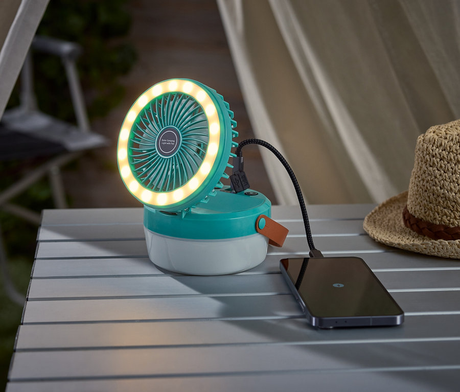 Ein türkisfarbener Campingventilator mit Lichtring lädt ein Smartphone auf einem Campingtisch auf, neben einem Strohhut.