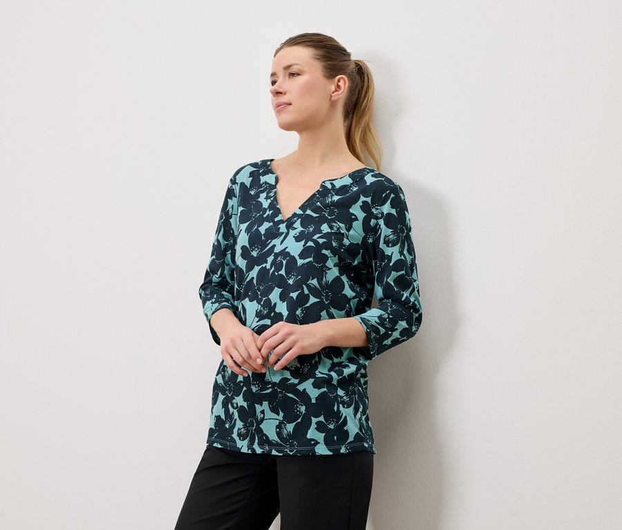 Frau mit blonden Haaren trägt eine blaugrüne Bluse mit Blumendruck und schwarze Hose.