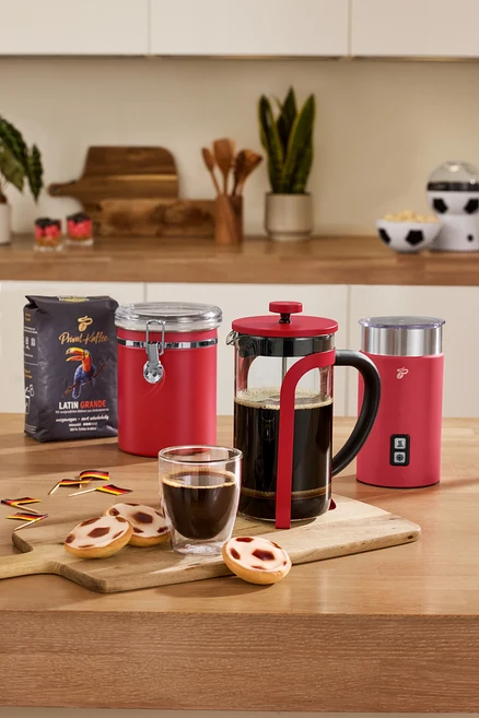 Auf einem Tisch stehen eine rote Aromadose, ein roter, elektrischer Milchaufschäumer, eine rote Siebstempelkanne, 2 Caffè Crema Gläser und eine Kaffeepackung.