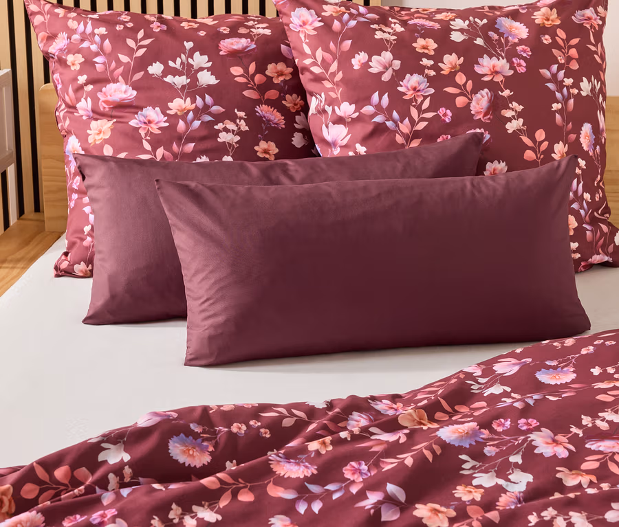 Auf einem Bett liegen zwei burgunderfarbene Kissen und zwei Kopfkissen mit Blumenmuster. Eine burgunderfarbene Bettdecke liegt am Fußende.