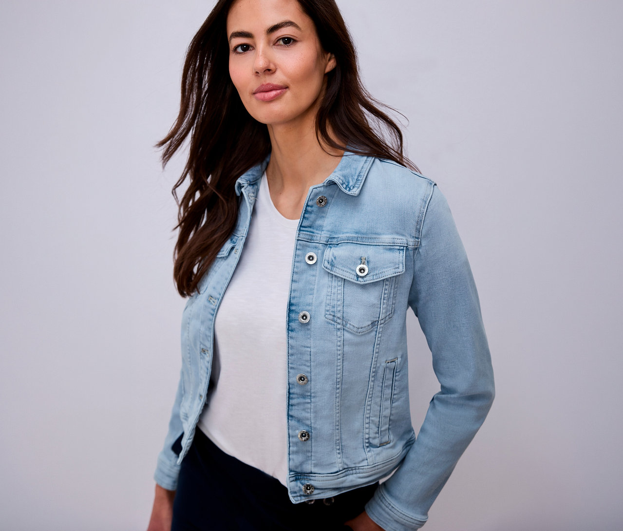 Frau mit braunen Haaren, die eine hellblaue Jeansjacke über einem weißen T-Shirt trägt.