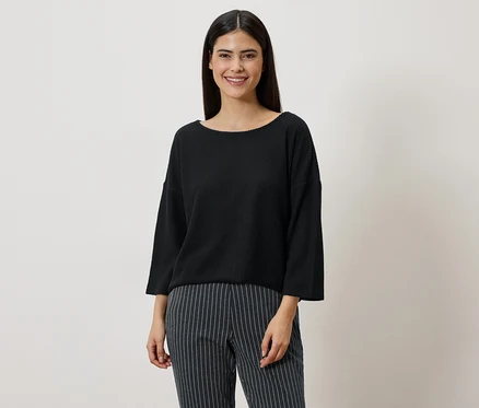 Frau mit dunklen Haaren trägt einen schwarzen Pullover und eine gestreifte Hose vor einem neutralen Hintergrund.