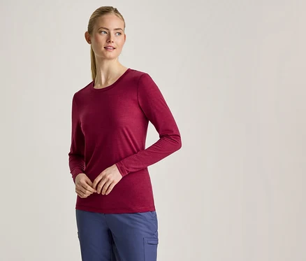Eine Frau mit blondem Pferdeschwanz trägt ein langärmeliges, weinrotes Shirt und blaue Hosen, steht mit vor dem Körper verschränkten Händen.