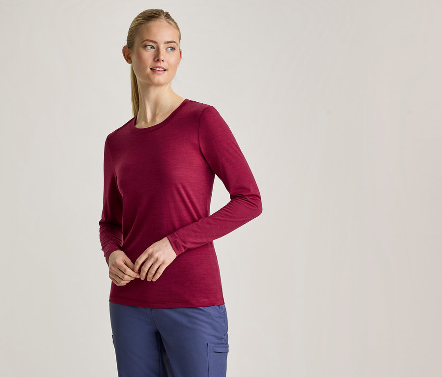 Eine Frau mit blondem Pferdeschwanz trägt ein langärmeliges, weinrotes Shirt und blaue Hosen, steht mit vor dem Körper verschränkten Händen.