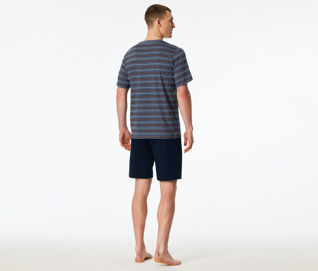 Ein Mann von hinten, der ein gestreiftes SCHIESSER Shorty-Set mit blauem, braunem und grauem Oberteil sowie marineblauen Shorts trägt. Er steht barfuß.