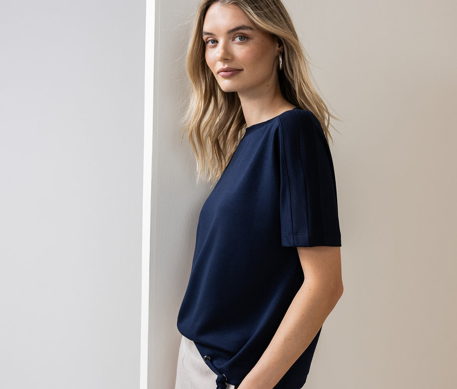 Frau lehnt an einer Wand und trägt ein marineblaues T-Shirt und eine helle Hose.