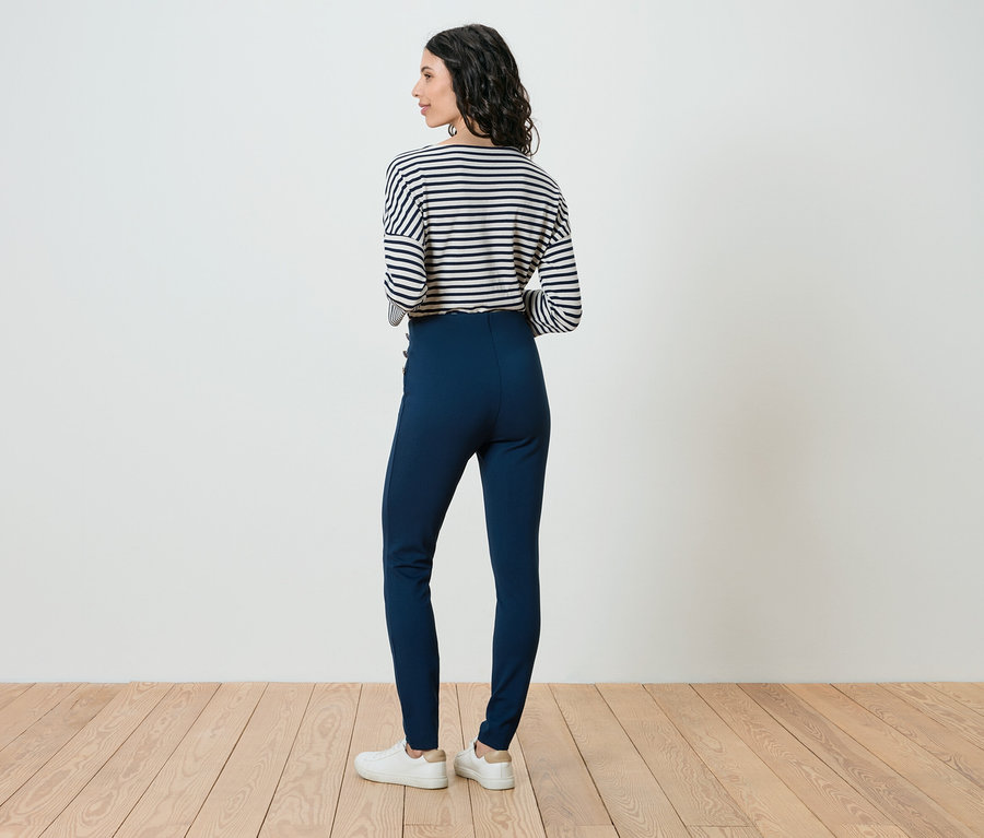 Frau steht mit dem Rücken zur Kamera, trägt ein gestreiftes Oberteil, blaue Hose und weiße Schuhe.