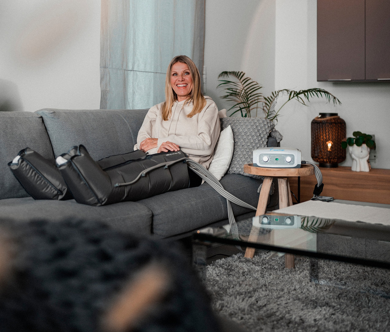 Frau entspannt mit dem Venen Engel 4 Gleitwellenmassage-Gerät mit Beinmanschette auf einem grauen Sofa im Wohnzimmer.