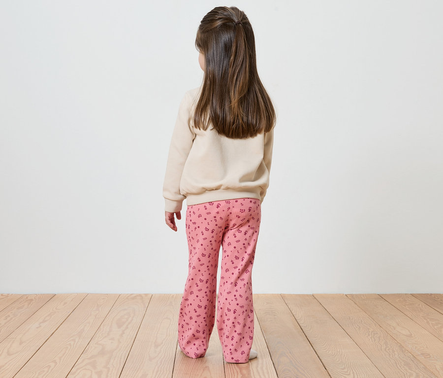 Rückansicht eines Mädchens mit braunen Haaren, beigem Pullover und rosafarbener Hose mit Blumenmuster.