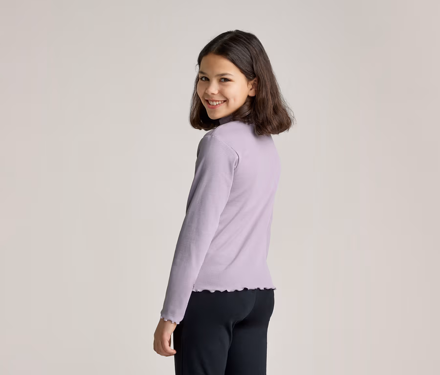 Ein junges Mädchen mit dunklen Haaren, das ein hellviolettes Langarmshirt mit Stehkragen und Rüschen an Ärmeln und Saum trägt, dreht den Kopf, um in die Kamera zu lächeln. Sie trägt auch eine schwarze Hose.