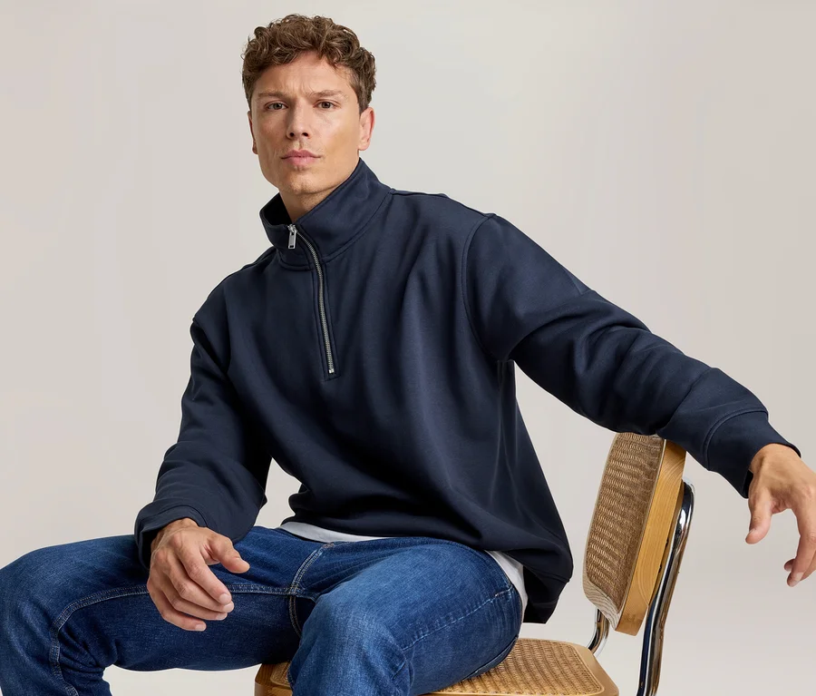 Mann sitzt auf einem Stuhl und trägt ein dunkelblaues Sweatshirt mit Reißverschluss und blaue Jeans.