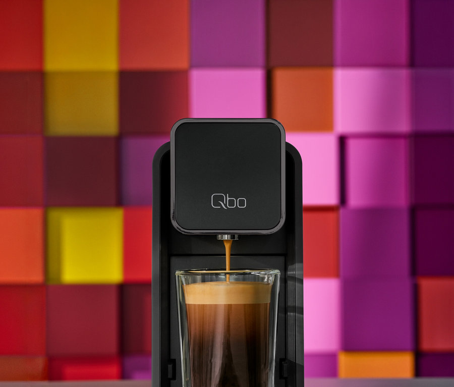 Schwarze Qbo Kaffeemaschine füllt Kaffee in ein klares Glas. Im Hintergrund eine bunte Wand mit quadratischen Fliesen in Rot-, Orange-, Gelb-, Pink- und Violetttönen.