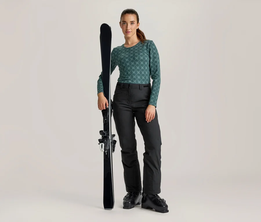 Frau steht, hält einen schwarzen Ski. Sie trägt ein grünes, gemustertes Langarmshirt, schwarze Skihosen und schwarze Skischuhe.