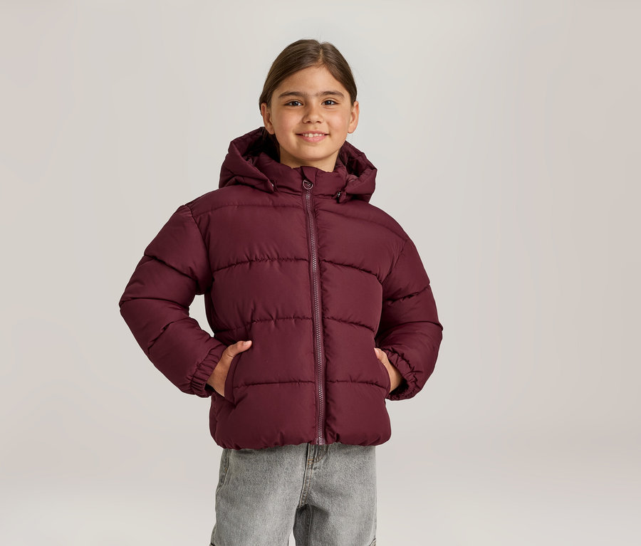 Lächelndes Mädchen in bordeauxroter Steppjacke mit Kapuze und grauer Jeans, die Hände in den Jackentaschen.