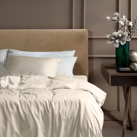 Ein Bett mit beigem Kopfteil und perlmuttfarbener SCHIESSER Satin-Bettwäsche "ELLA" sowie einem passenden Kissenbezug. Rechts steht ein Nachttisch mit einer grünen Vase und Dekoration.