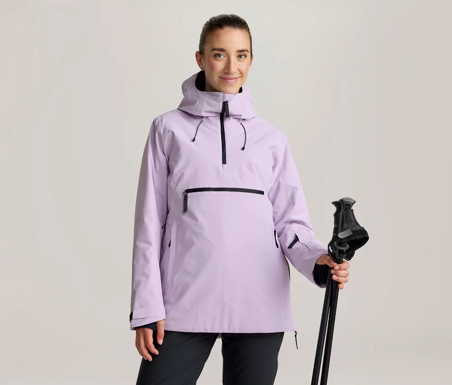 Frau in einer hellvioletten Anorak-Jacke mit schwarzen Reißverschlüssen und schwarzer Hose, die schwarze Skistöcke hält.