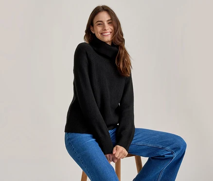 Lächelnde Frau mit langen braunen Haaren, die einen schwarzen Rollkragenpullover und blaue Jeans trägt und auf einem Holzhocker sitzt.